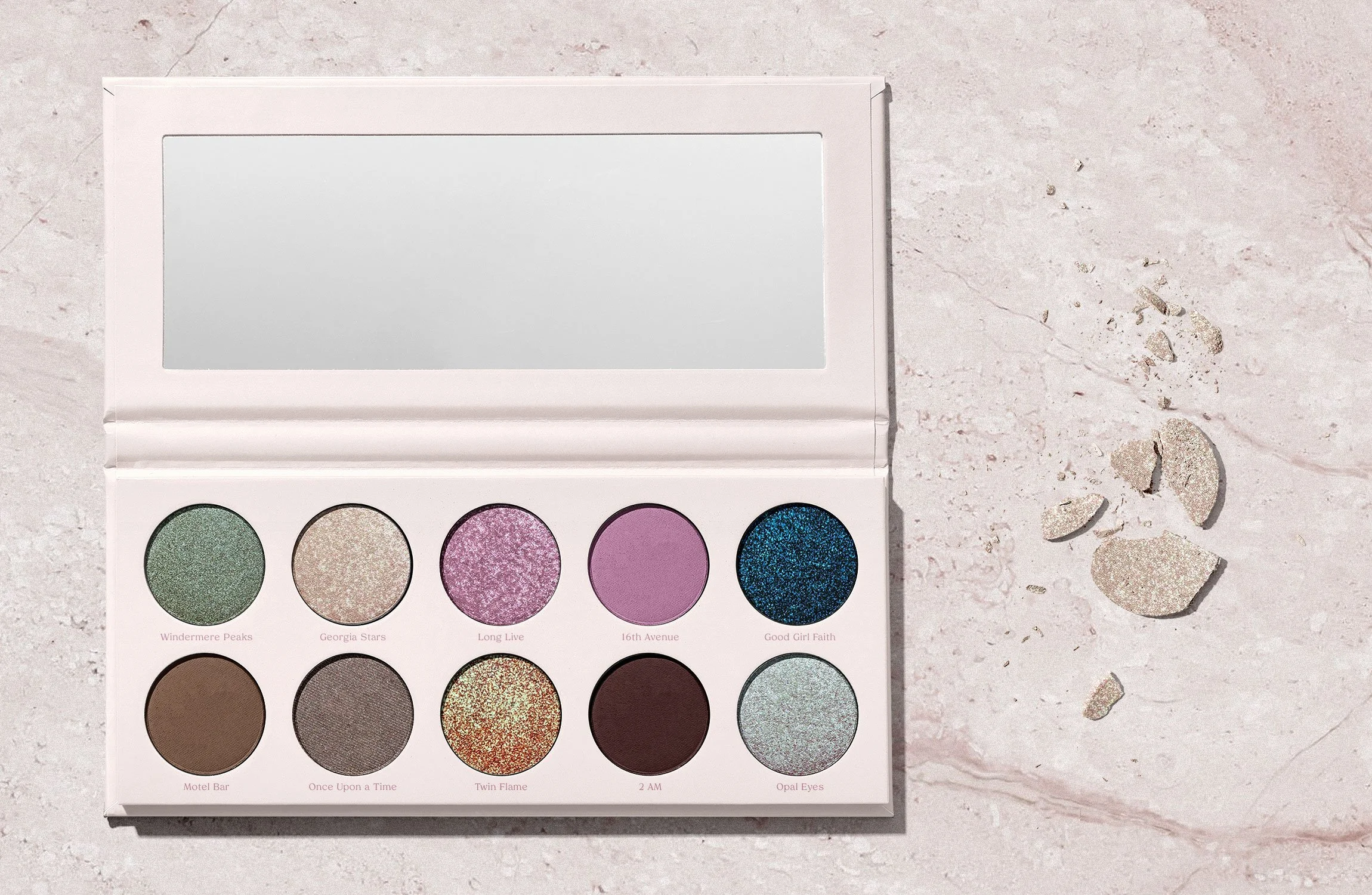 Open eyeshadow palette with nine shades and a mirror, placed on a light pink marble surface with some broken chunks of material nearby.