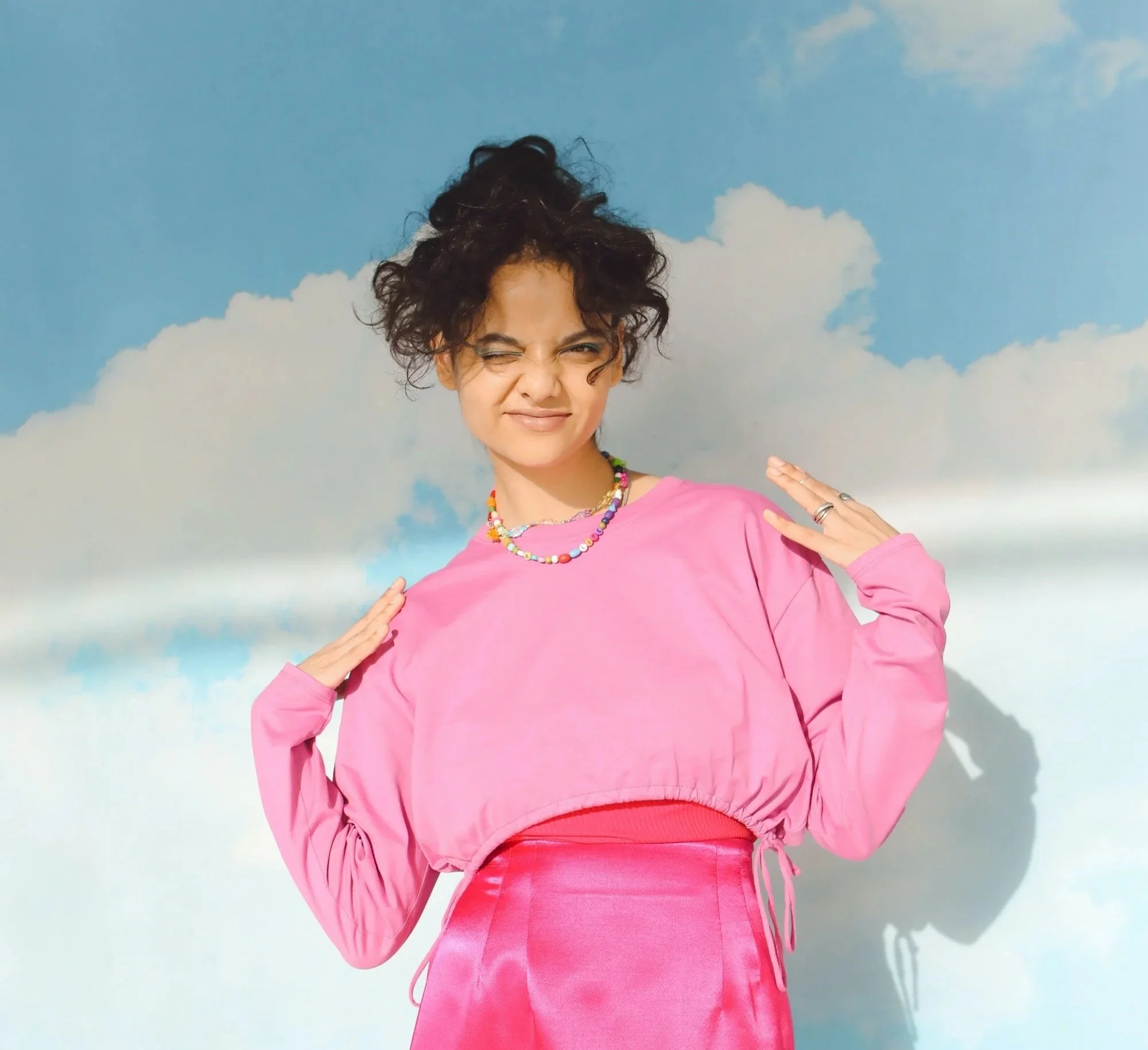 Young woman in pink outfit making a playful expression against a cloudy sky background.
