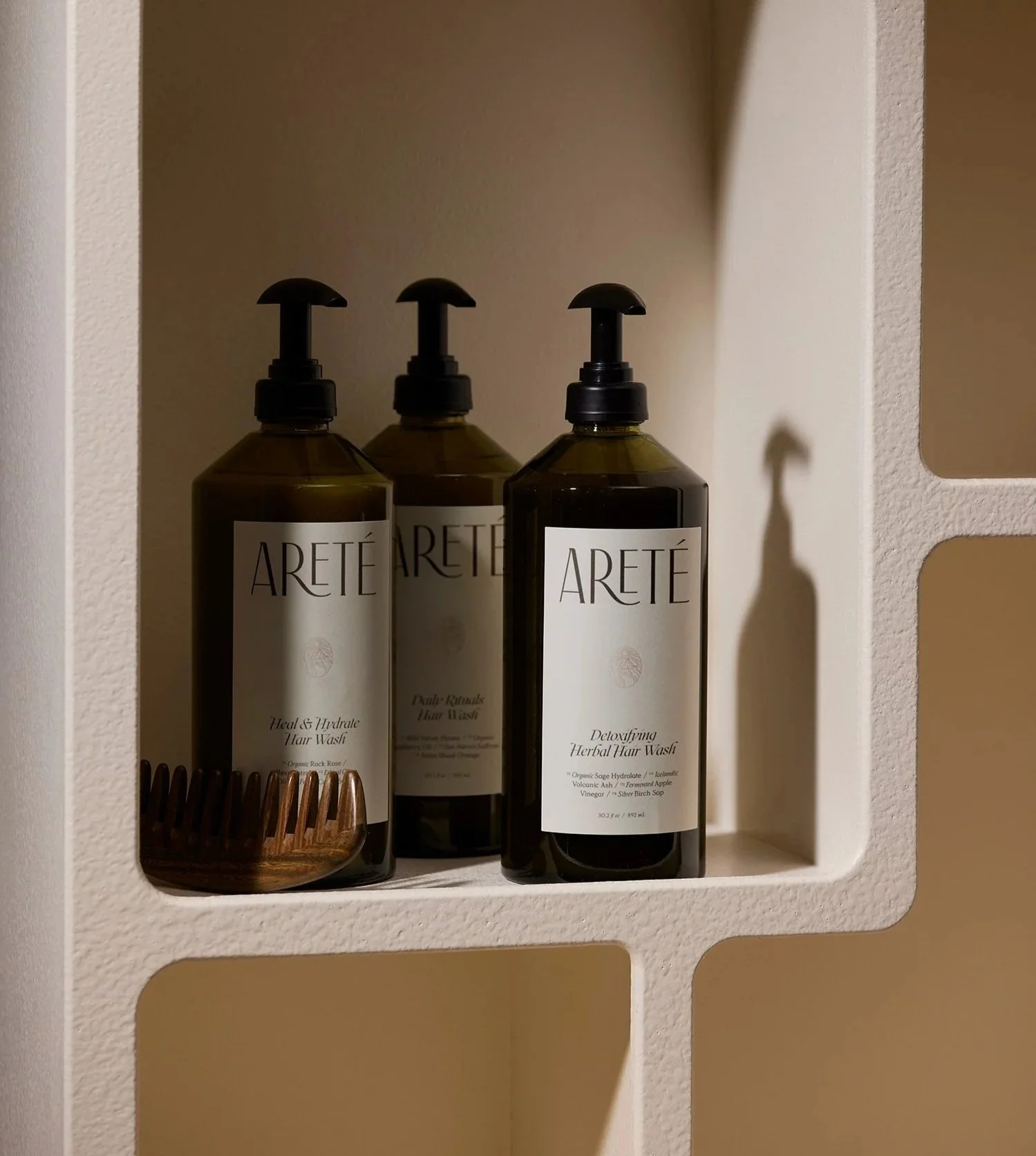 Three dark-colored bottles of hair wash with pump dispensers on a white shelf, labeled 'ARETÉ', in a shower niche.