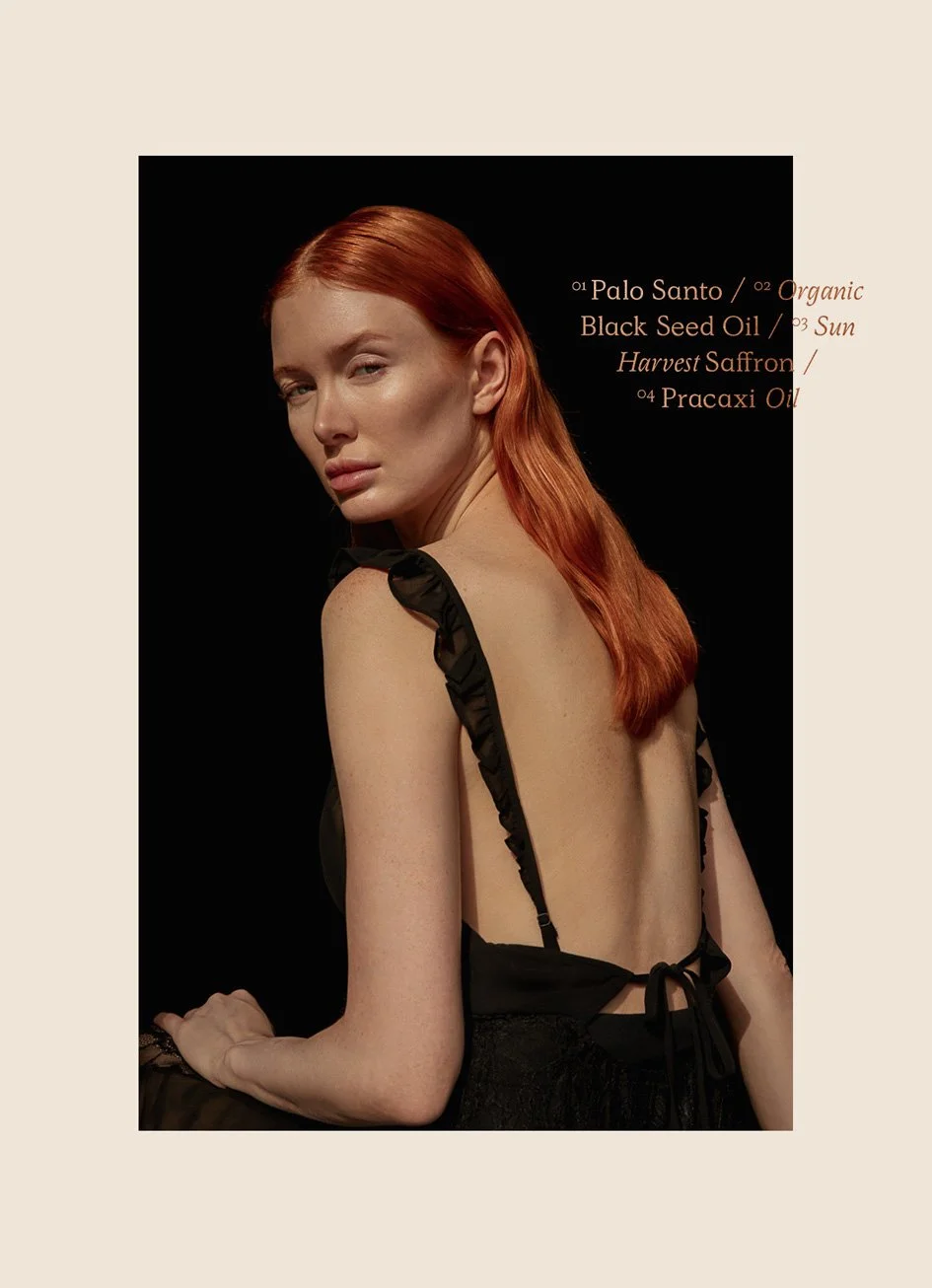 A woman with red hair sitting against a black background, wearing a black backless top with ruffled straps, and text listing ingredients such as Palo Santo, Black Seed Oil, Sun Harvest Saffron, and Pracaxi Oil.