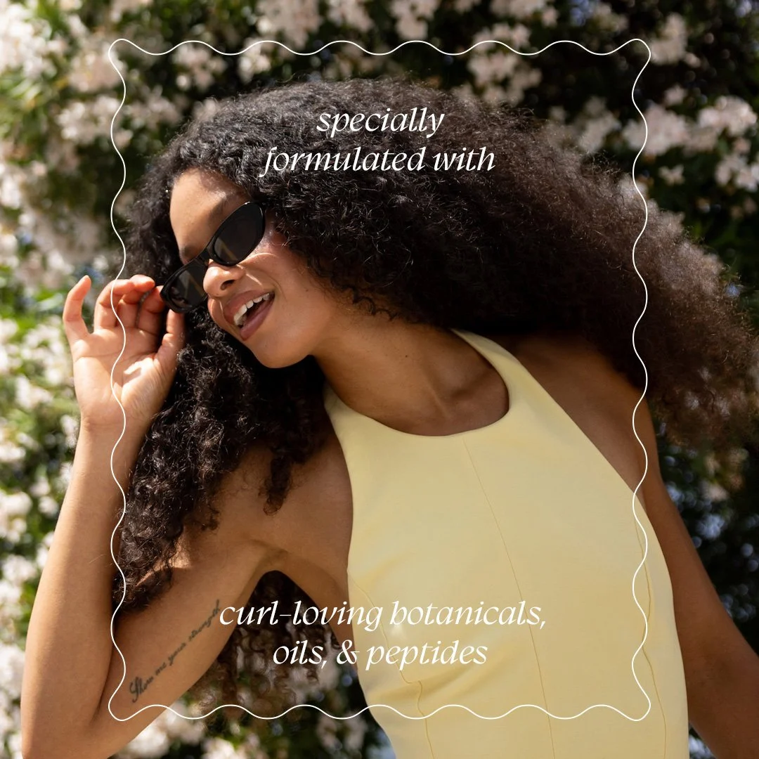A woman with curly hair wearing sunglasses and a yellow top outdoors, smiling with a background of blooming bushes. Text overlays say 'specially formulated with curl-loving botanicals, oils, & peptides.'