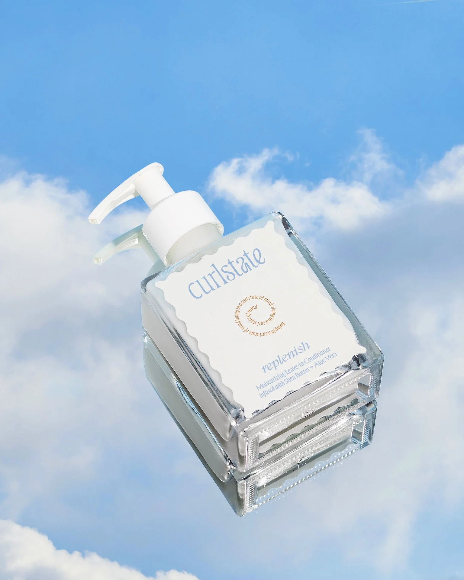 A clear plastic bottle of Curlstate replenish moisturizing leave-in conditioner with a white pump dispenser, floating in a sky with clouds.