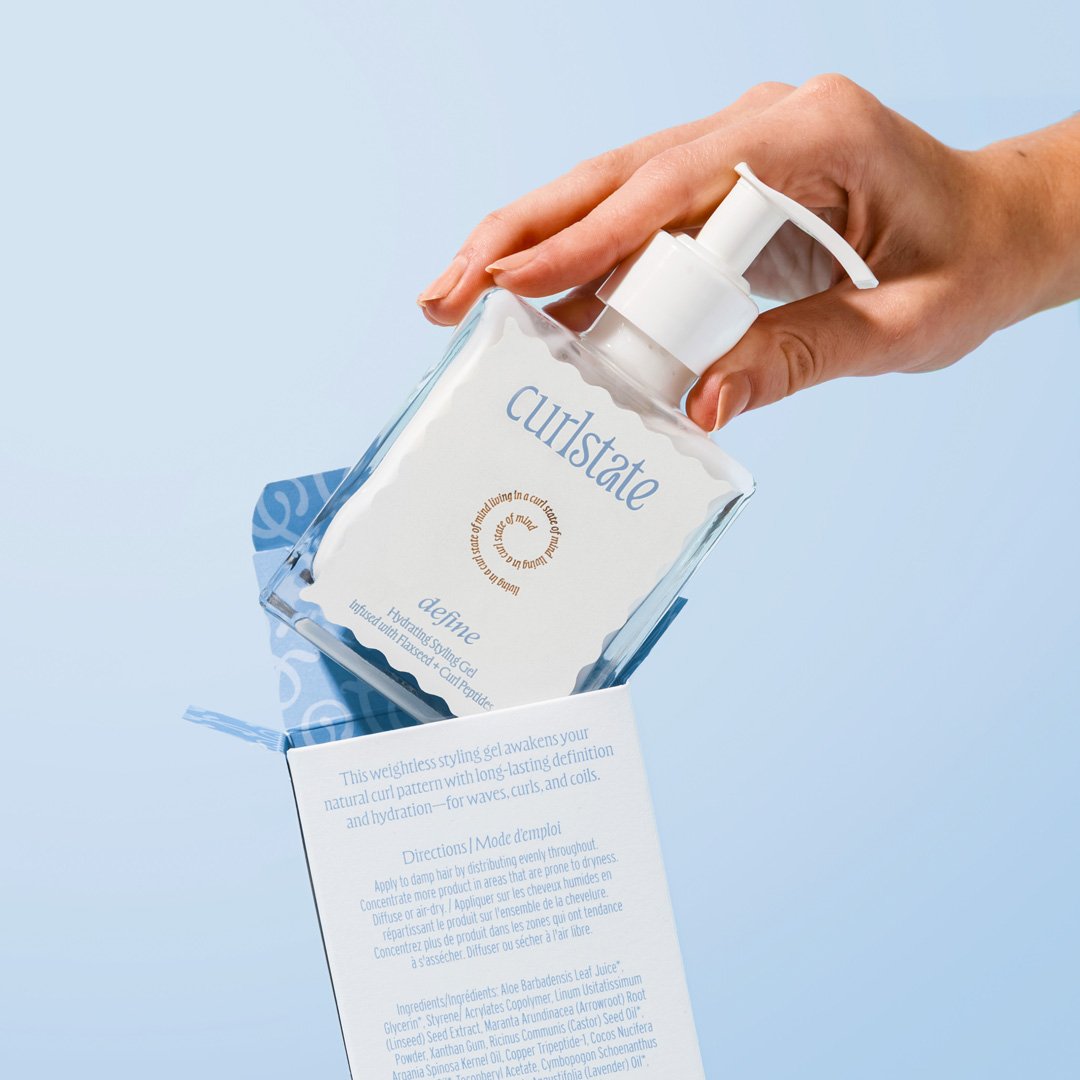 A hand holding a clear plastic bottle of Curlstate define hydrating styling gel against a light blue background, with a product label visible.