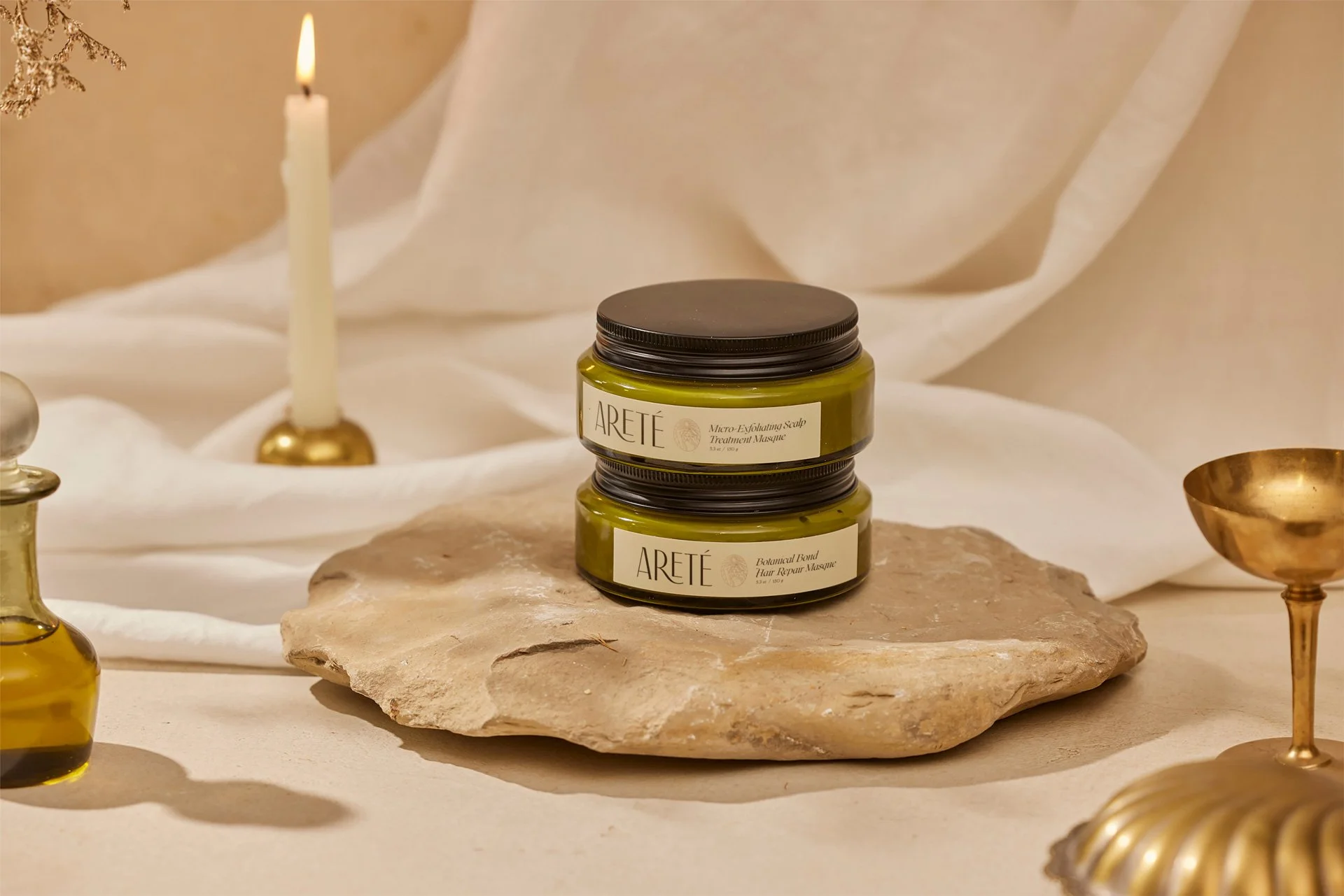 Two green jars labeled Arete, stacked on a large beige stone in a beige and white setting, surrounded by gold-colored objects including a candle, a small bottle, and a bowl.