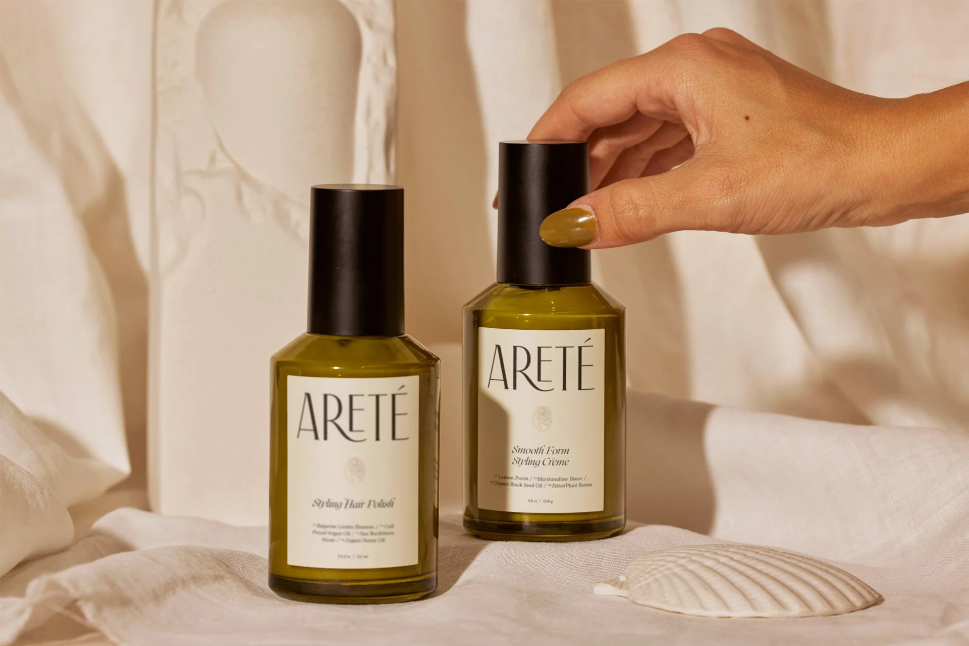 Two bottles of ARETÉ hair styling products on a white surface, with a hand adjusting the cap of one bottle, and a seashell nearby, against a neutral background.