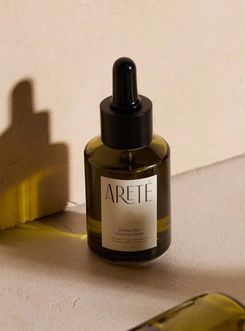 A dark green glass bottle with a black dropper cap labeled "ARETÉ" and "Healing Hair Ceremony Serum", sitting on a beige surface with a beige wall behind it. The shadow of the bottle is cast on the wall.