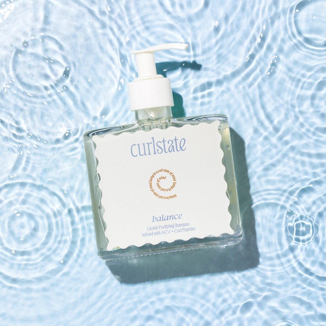 A clear bottle of curlstate balance gentle purifying shampoo with a white pump, floating on water with ripples.