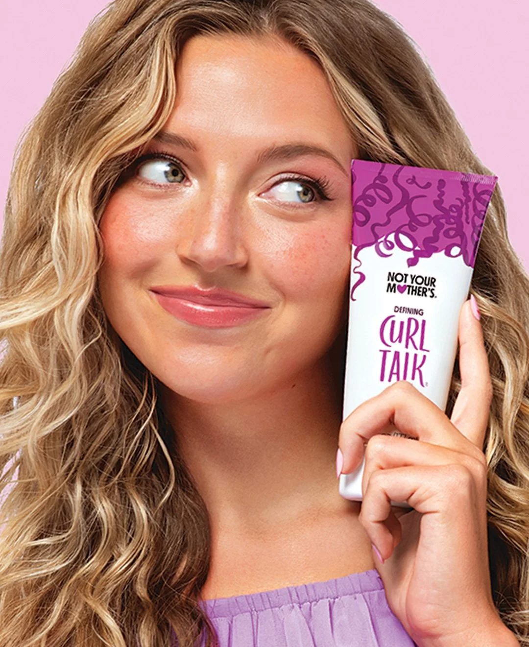 A smiling woman with curly blonde hair holding a tube of Not Your Mother's Curl Talk hair product, against a pink background.