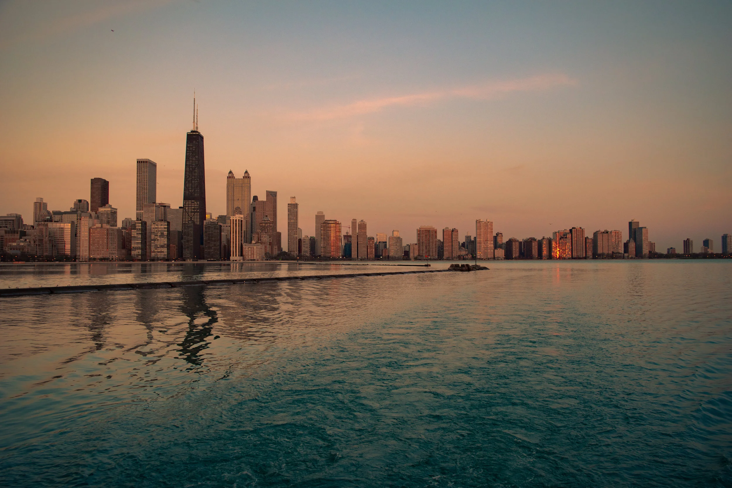 Chicago skyline at sunset with tall buildings, including the Willis Tower, reflected on Lake Michigan.