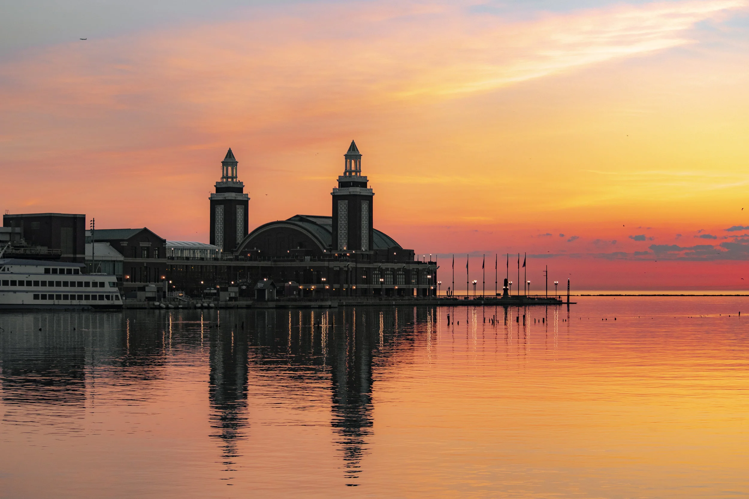 Sunset over a waterfront with a large building featuring two tall towers and a curved roof, reflected in the calm water.