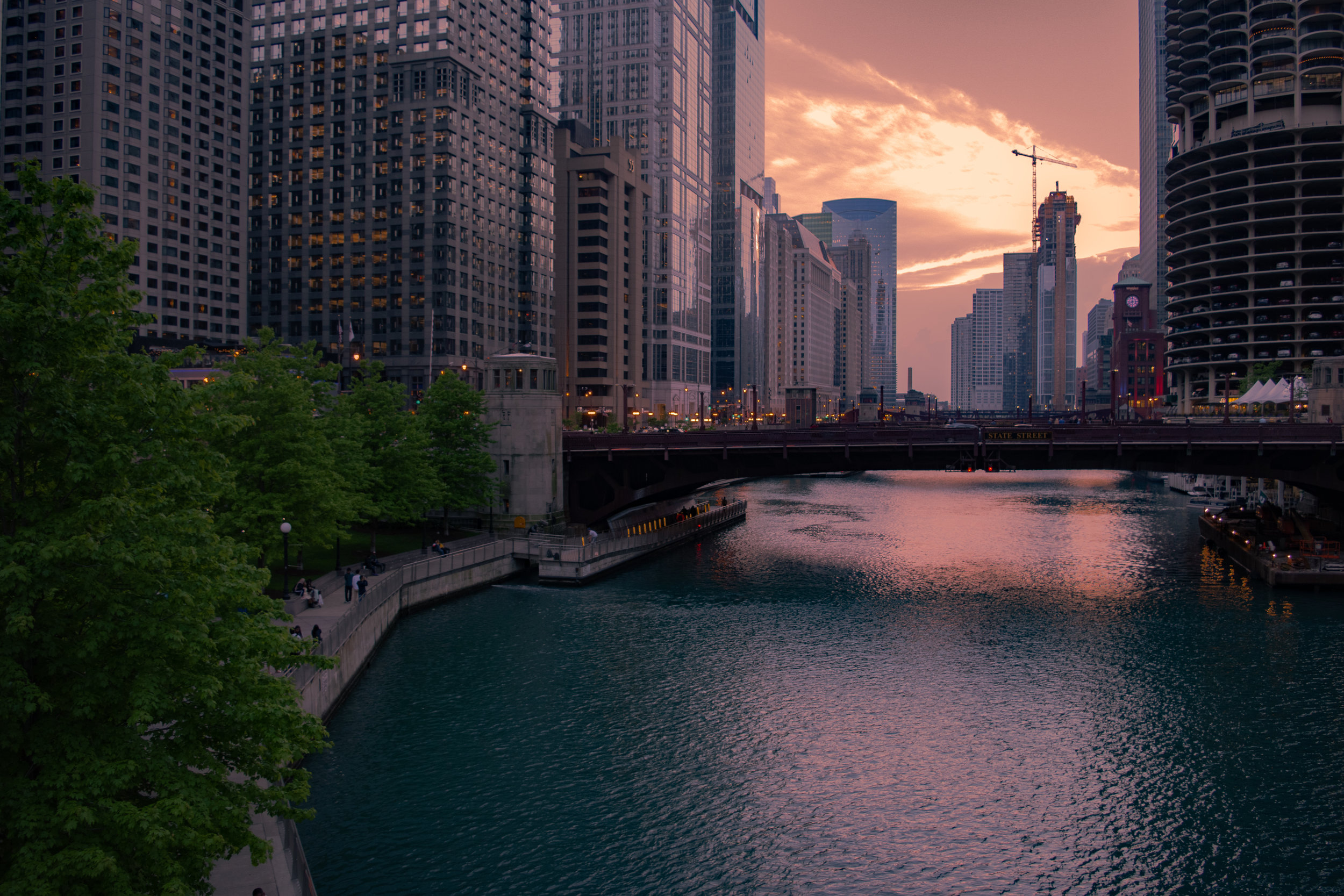 View of a city skyline with tall buildings along a river at sunset, with a bridge over the river and a promenade on the left side with trees and people.