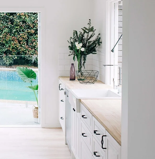 Three Birds Renovations House 2 kitchen with white cabinetry, timber benchtop, farmhouse sink and fresh greenery overlooking poolside outlook.