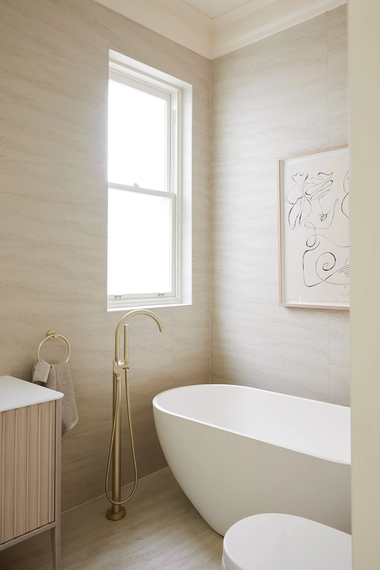  Three Birds Renovations House 17 bathroom featuring a freestanding bath beneath a window, brushed brass tapware, stone-look wall tiles and a fluted vanity. 