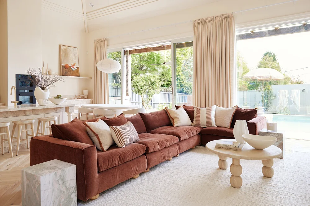 Three Birds Renovations House 17, warm open-plan living room with rust velvet sectional, round timber coffee table, and pool views, blending texture and light for relaxed contemporary living.