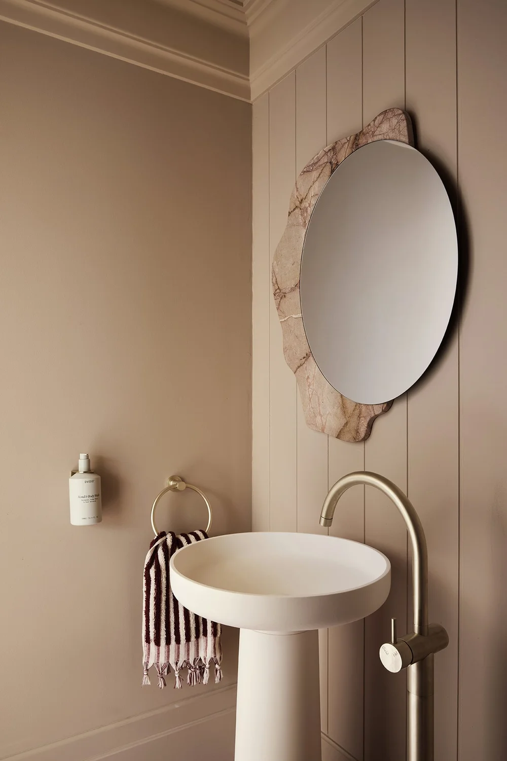 Three Birds Renovations House 17, stylish powder room with pedestal basin, round stone-edged mirror, brass tapware, and warm taupe walls creating a sophisticated modern aesthetic.