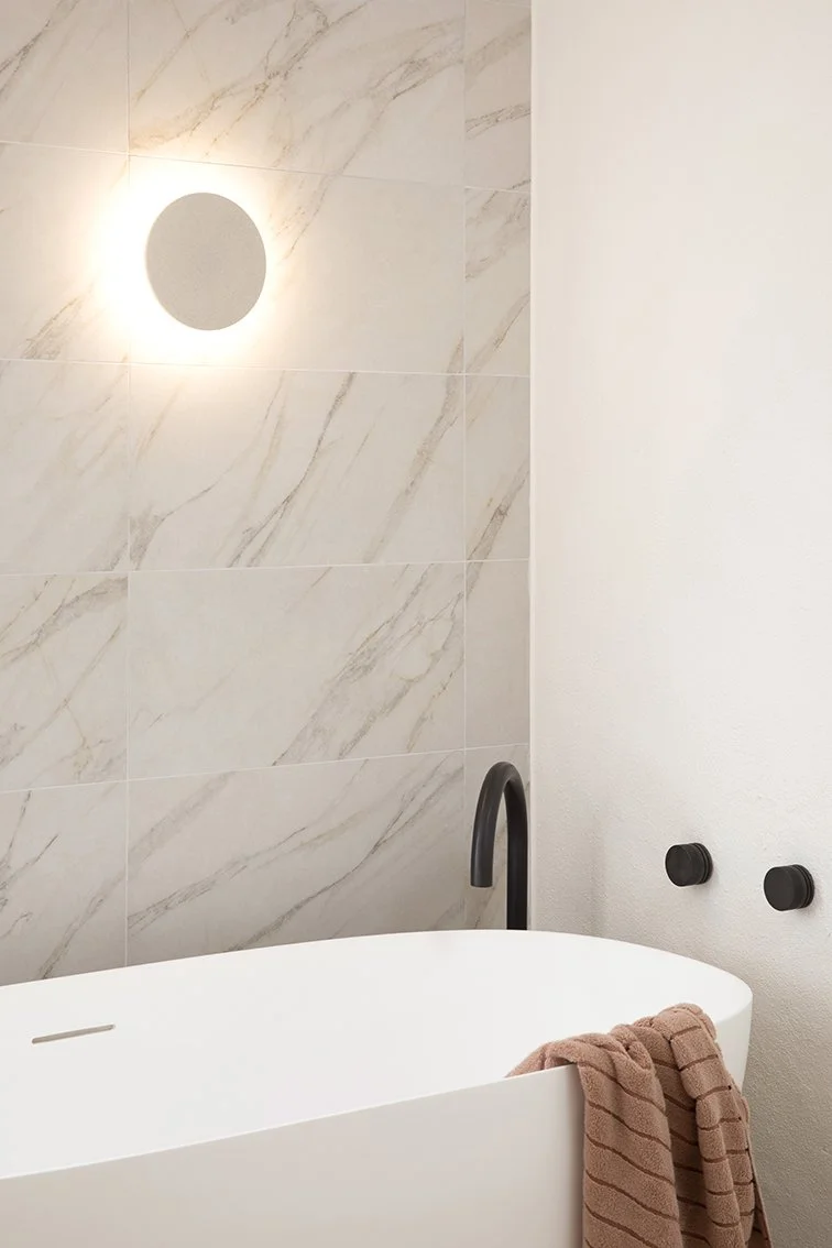 Three Birds Renovations House 15 bathroom with a freestanding bath, marble-look wall tiles, matte black tapware and soft ambient lighting.