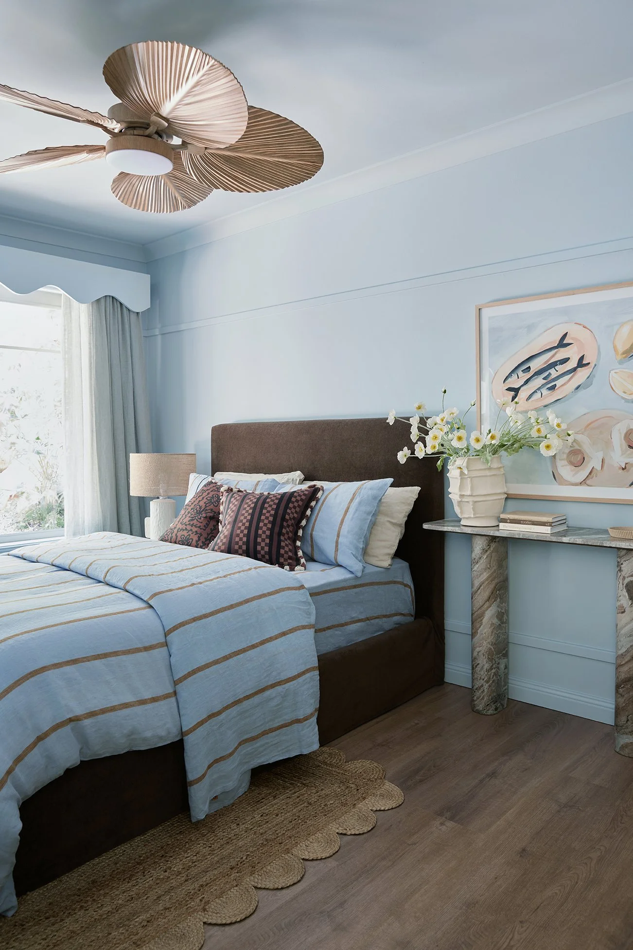 Three Birds Renovations Eclectic Escape bedroom with sky blue walls, upholstered bedhead, striped linen bedding and styled bedside vignette with artwork and fresh florals.