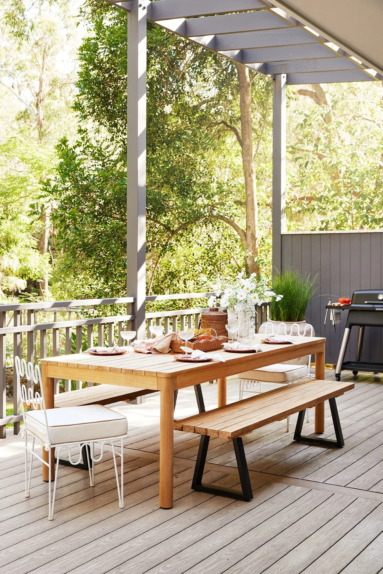 Three Birds Renovations Eclectic Escape outdoor dining deck with timber table, bench seating and leafy bushland backdrop.