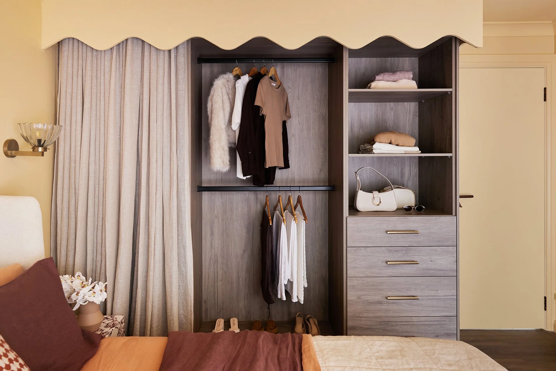 Three Birds Renovations Eclectic Escape bedroom with open wardrobe, timber-look cabinetry, brass handles and scalloped pelmet over soft linen curtains.