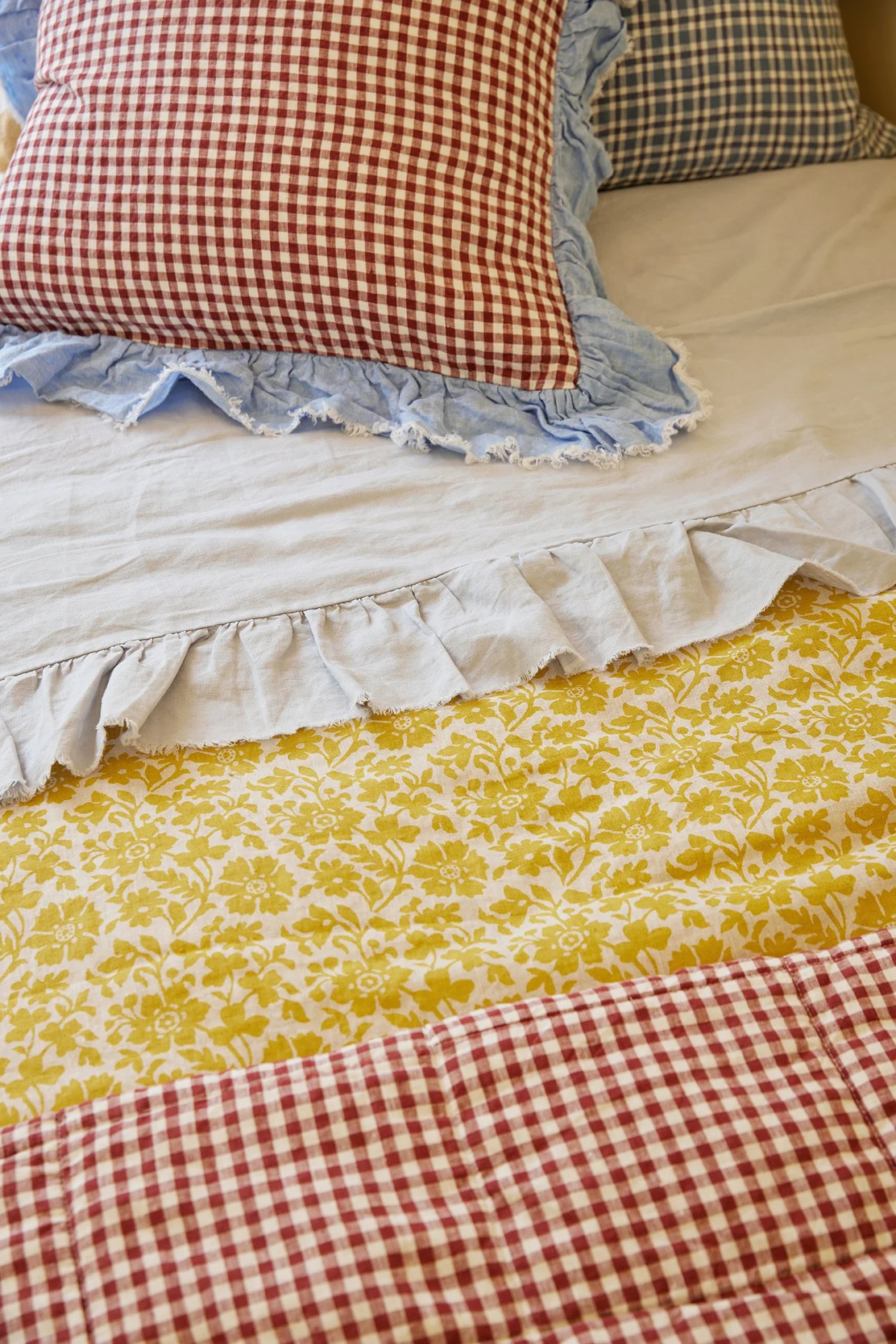 Three Birds Renovations Eclectic Escape layered bed styling with yellow floral sheets, red gingham quilt and soft blue ruffle pillows.