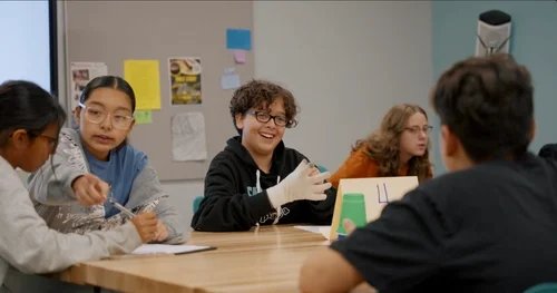 Students collaborating and smiling at a table in a bright classroom setting for learning content.