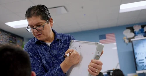 Teacher in glasses writing a math equation on a small whiteboard in a classroom setting.