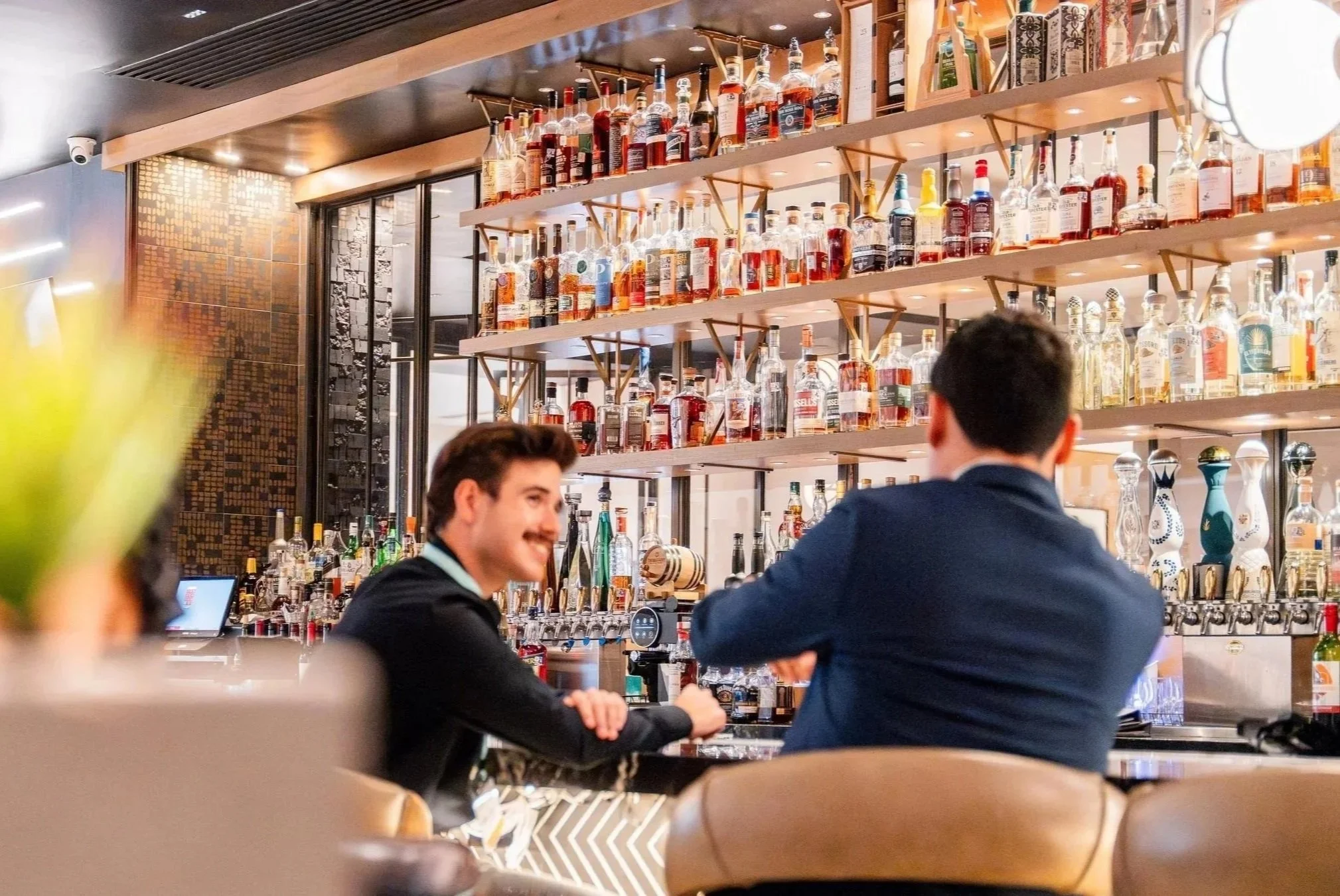 Bar host serving a guest at an upscale hotel bar with backlit liquor shelves for hospitality commercial photography.