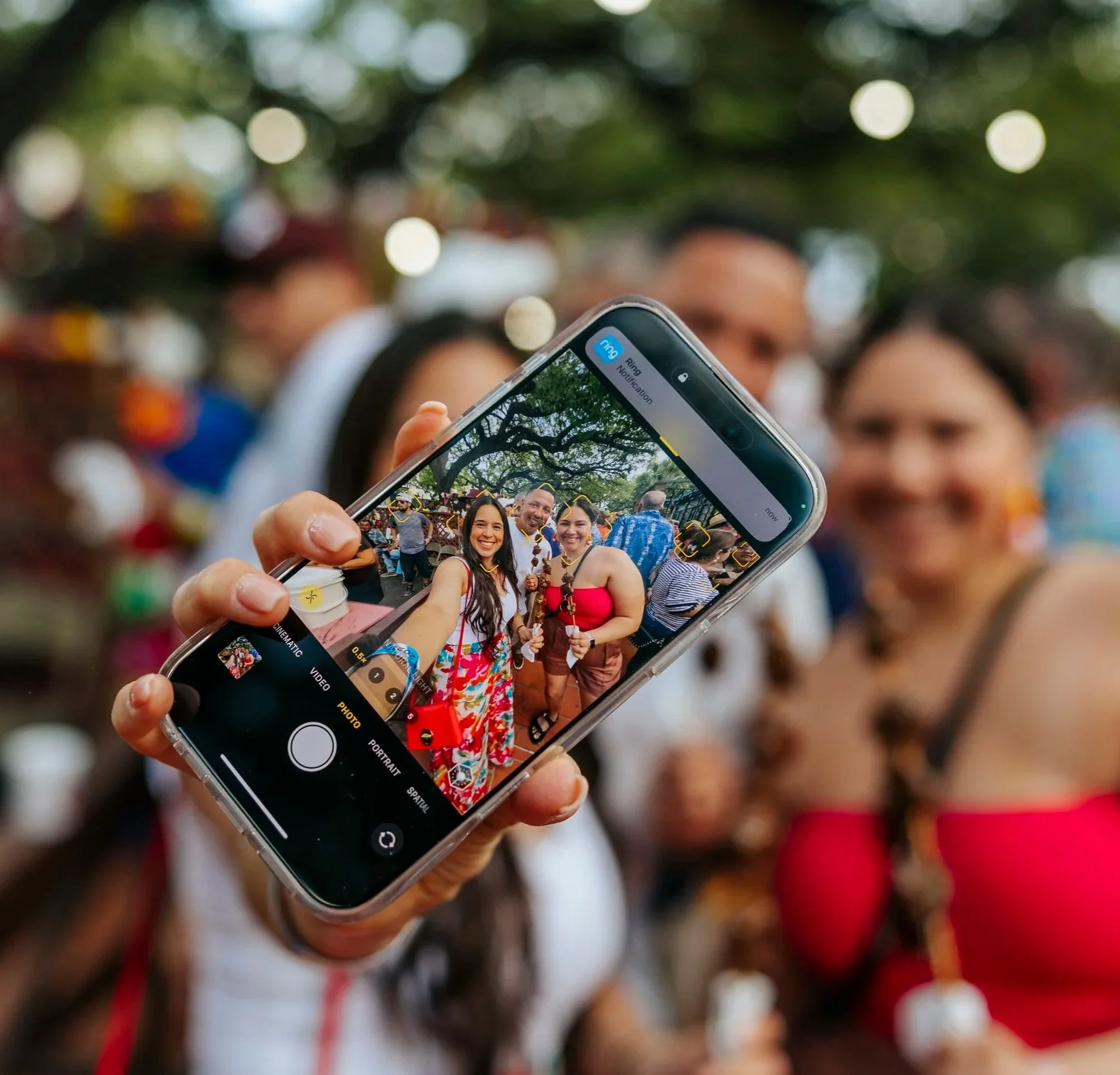 Person holding up a phone showing a group selfie at a colorful outdoor San Antonio festival.