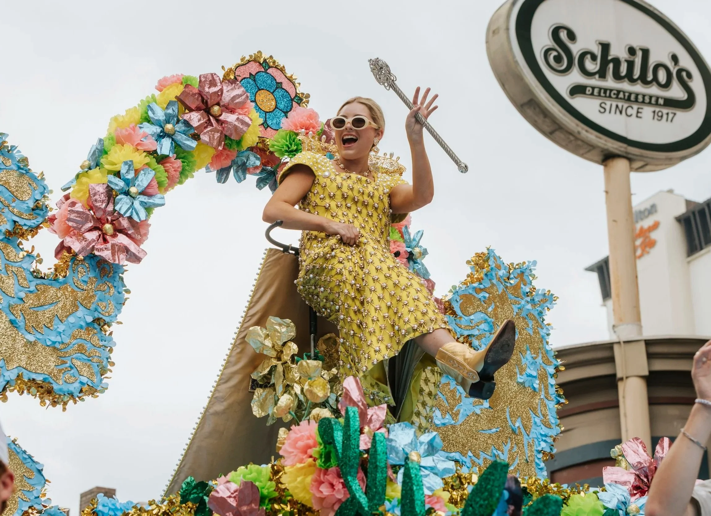 Smiling woman in a yellow gown waving from a colorful float during San Antonio Fiesta parade.