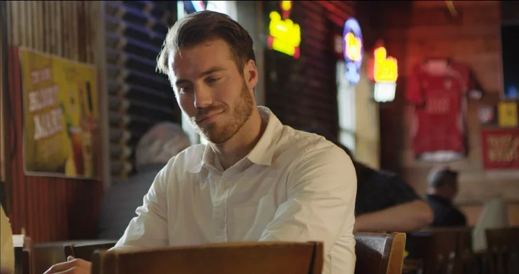Kyle Park sitting at a table in a bar setting, a scene from the Don't Forget Where You Come From video.