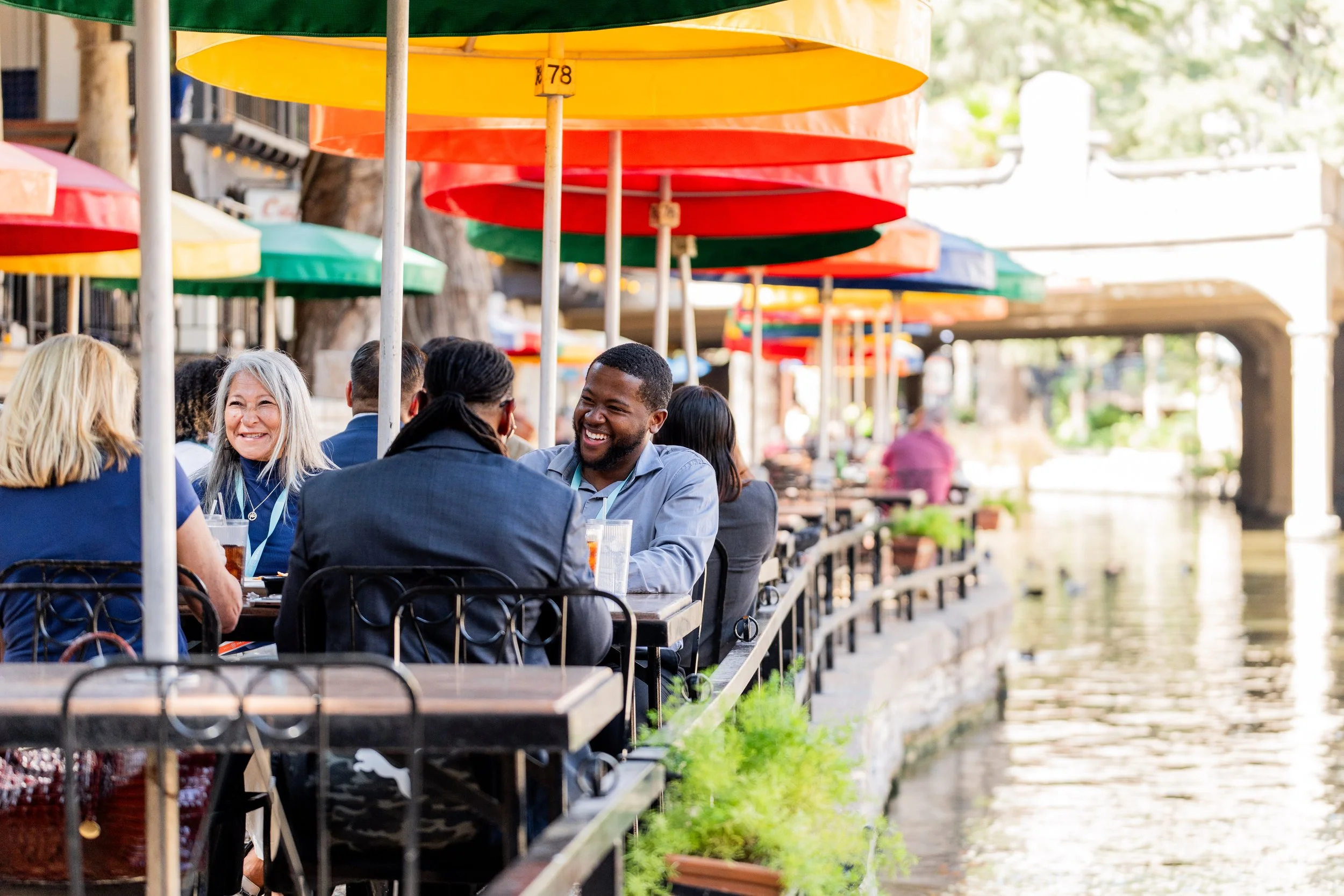 Group of professionals laughing and dining outdoors along the San Antonio River Walk.