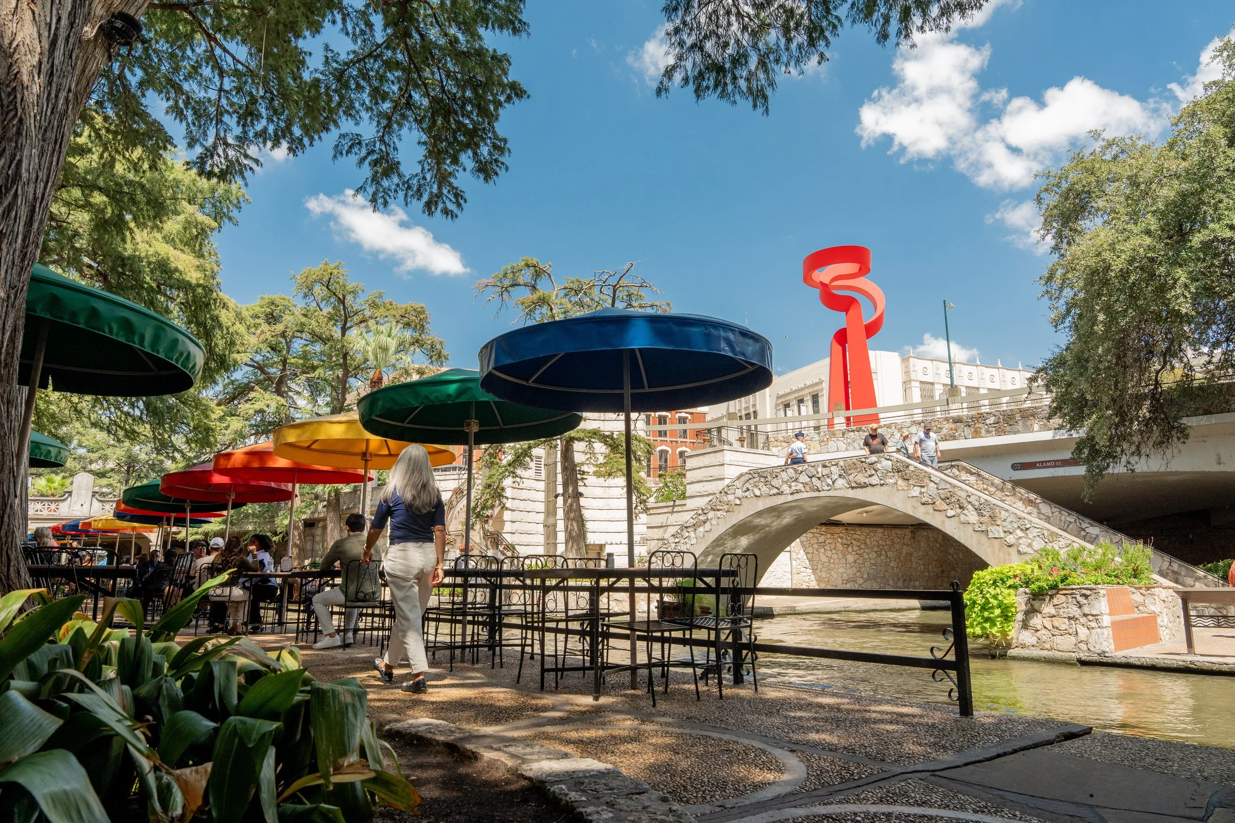 Colorful outdoor dining umbrellas and stone bridge along the San Antonio River Walk on a sunny day.