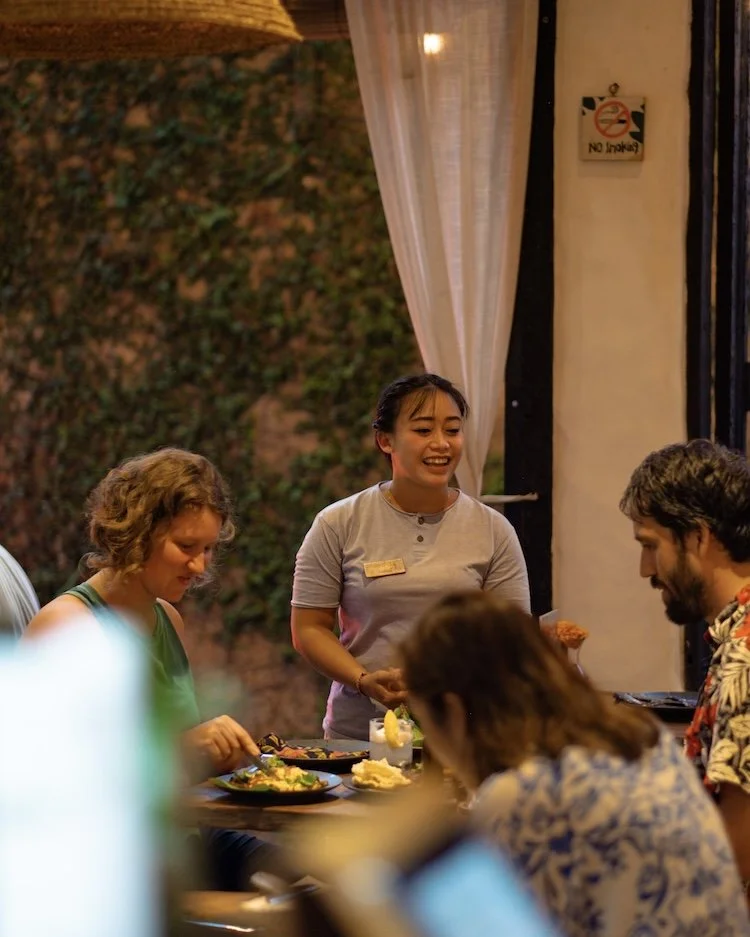 A smiling waitress with a name tag serving food to diners at a restaurant table with a leafy background and white curtains.