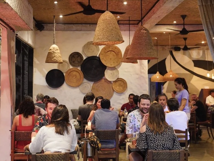 People dining at a Ubud's Best Indonesian restaurant with woven pendant lights and an art wall with circular wooden and black accents.