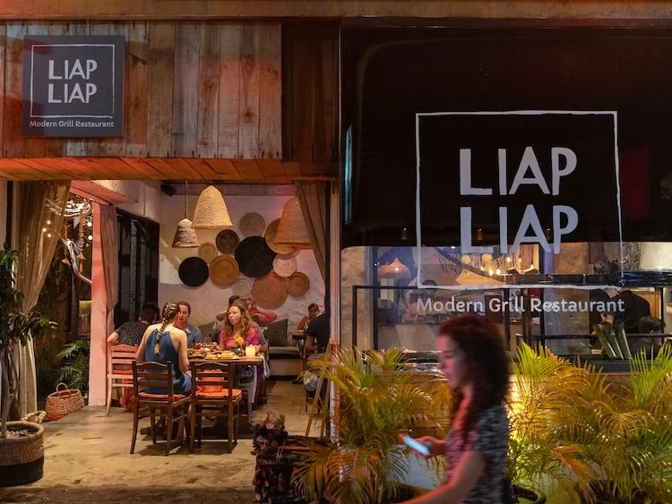 Exterior view of Liap Liap Best Modern Indonesian Grill Restaurant with outdoor seating, people dining, and decorative woven wall art inside