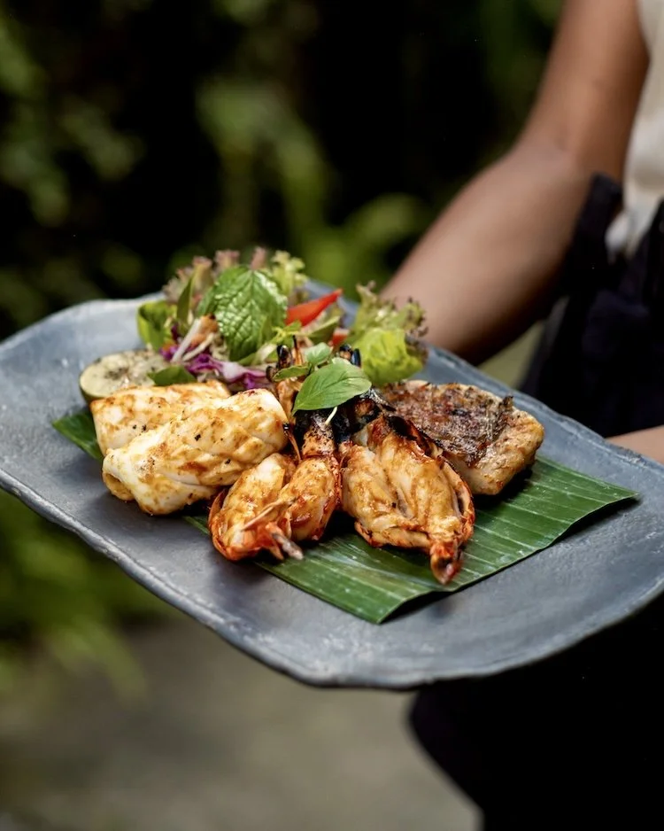 A black plate with grilled seafood and salad on a banana leaf, held by a person outdoors.