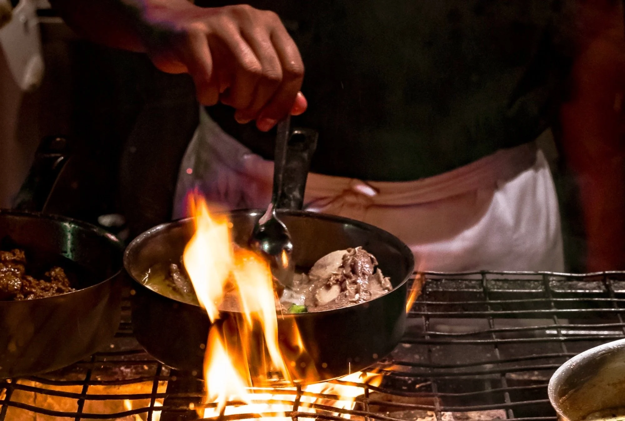 A person cooking with flames rising from a pan on a grill, holding a spoon over cooked meat in a black bowl, with other dishes nearby at Ubud's Best Indonesian restaurant.