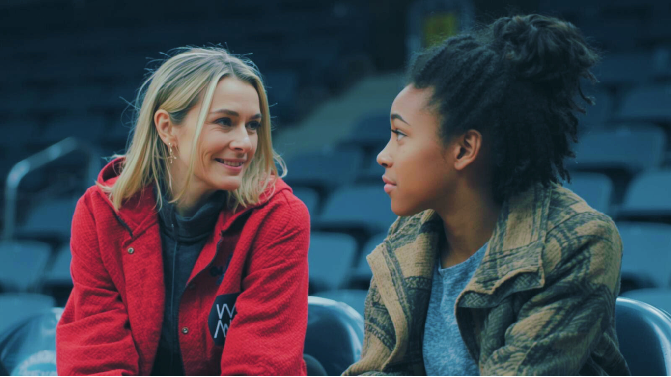 Cinematic slow-motion capture of a mentorship moment_ experienced female coach speaking with a young athlete courtside in Barcelona. Empty arena seats blurred behind them. Authentic emotional intensity. Real, unscripted body language