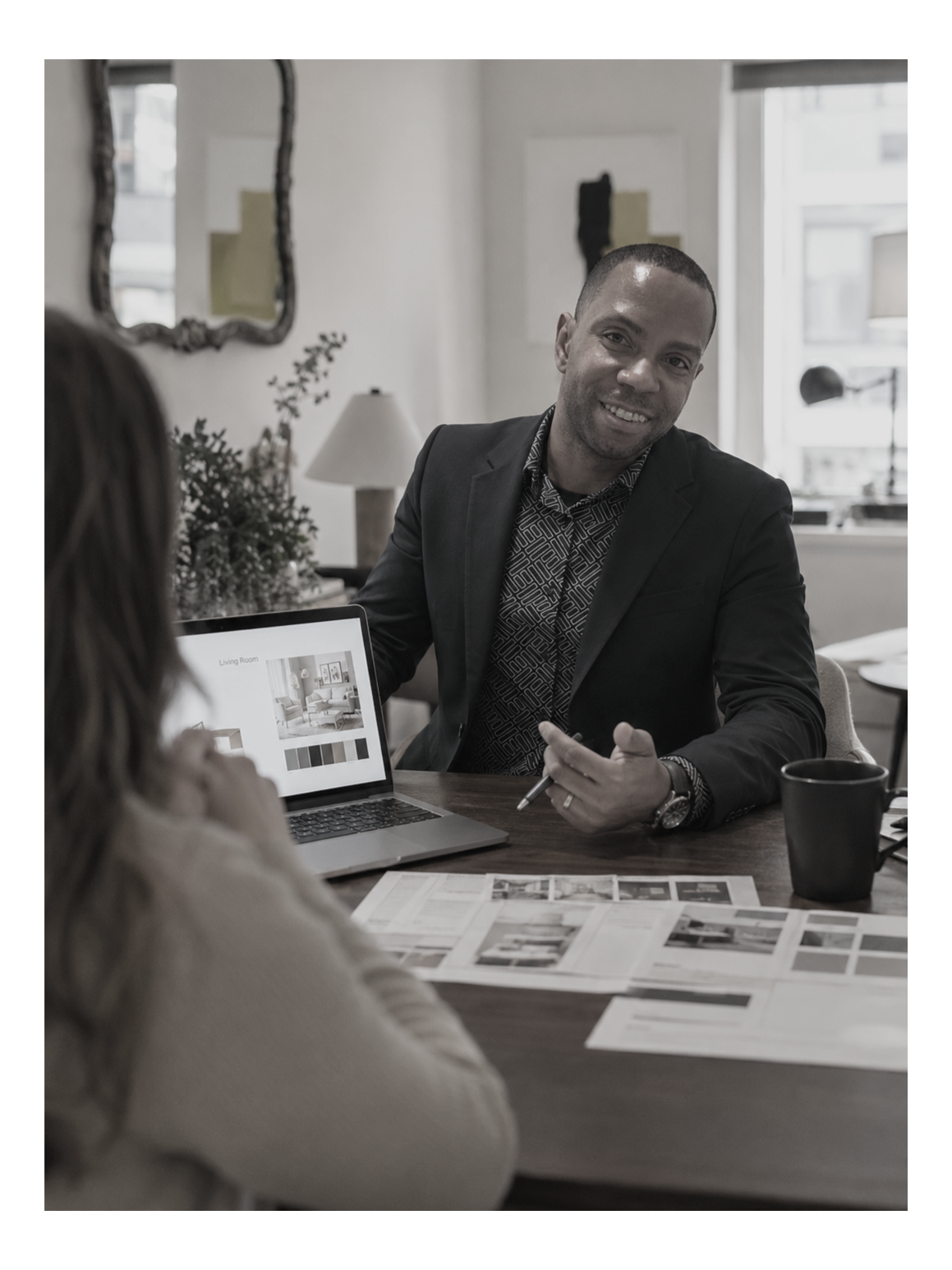 A man in a suit smiling during a conversation with a woman in an office or meeting room with a laptop, papers, and a cup on the table.