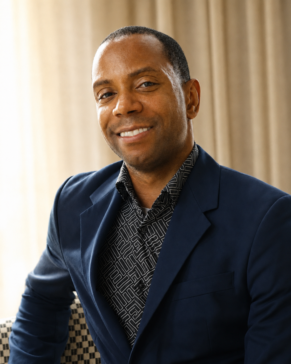 A smiling African American man in a navy suit and patterned shirt, indoors with beige curtains in the background.