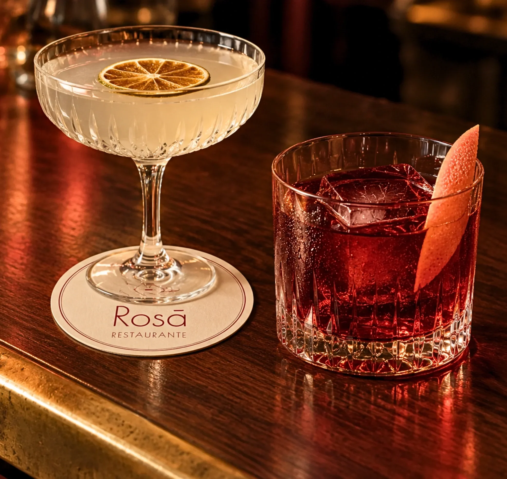 A cocktail glass with a lemon slice garnish on top and a red drink in a glass with ice and a lemon wedge on the side, on a dark wooden bar counter.