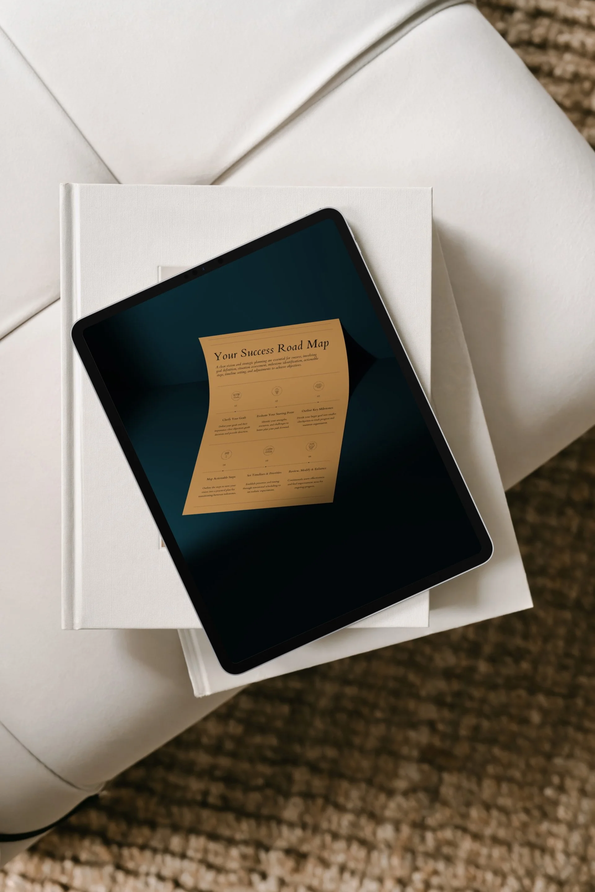 A tablet displaying a document titled 'Your Success Road Map' on top of two stacked books, resting on a white chair with a textured brown carpet underneath.
