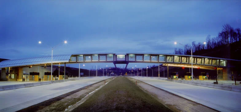 The Pennsylvania Turnpike Commission (PTC) desired a new toll plaza for the electronic fares of the future as well as a new image for their mainline facilities – one that would be beautiful as well as safe, cost effective, and adaptable. This facility was the first on the PTC system that would integrate high-speed toll collection as well as manned and unmanned booths. The manned booths will disappear in the “all electronic” future. This exciting architectural design features an innovation for the Turnpike: a bridge linking both sides of the Plaza, instead of the usual service tunnel. As a result of this bridge, the Plaza has a greater visual presence than is normally expected while acting as a new portal to the Pittsburgh area for traffic on the new Mon Fayette Expressway. EZ Pass readers are concealed in the architecture. This design is intended to set a new direction for future Turnpike Facilities. Design included a pedestrian bridge for access across the high-speed lanes, site development, lighting, tollbooths, canopies, signage, and utility building and appurtenances. Steel is the main element in the bridge and on the roof because of Pittsburgh’s history as the steel capital of the world. The facility was fully integrated with the highway system through many unique features that include: • Visual security techniques, gull winged canopies curved in two directions and an arched truss provide an exciting architecture. • A unique centrally located V-shaped pier supporting the “lookout” from which the all electronic plaza will be operated by one person. • Features that allow the toll plaza to be adaptable to the state-of-the-art Electronic Tolls and Traffic Management (ETTM) System. At the mainline facility, EZ Pass capabilities allow patrons to bypass the toll collection machines while maintaining a high rate of speed.