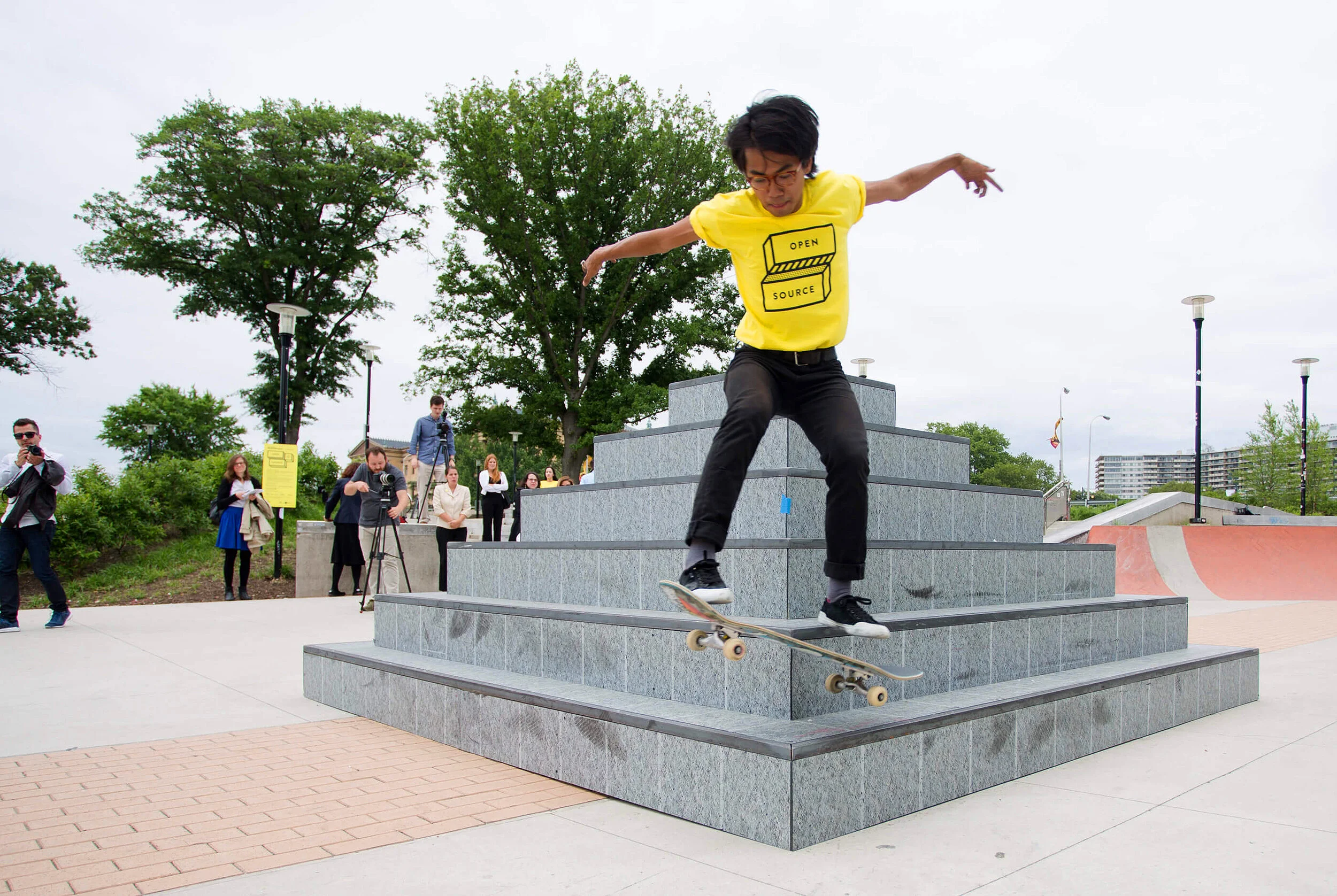 Photograph of a skater, wearing a yellow tshirt with a black Open Source logo, jumping off a structure.