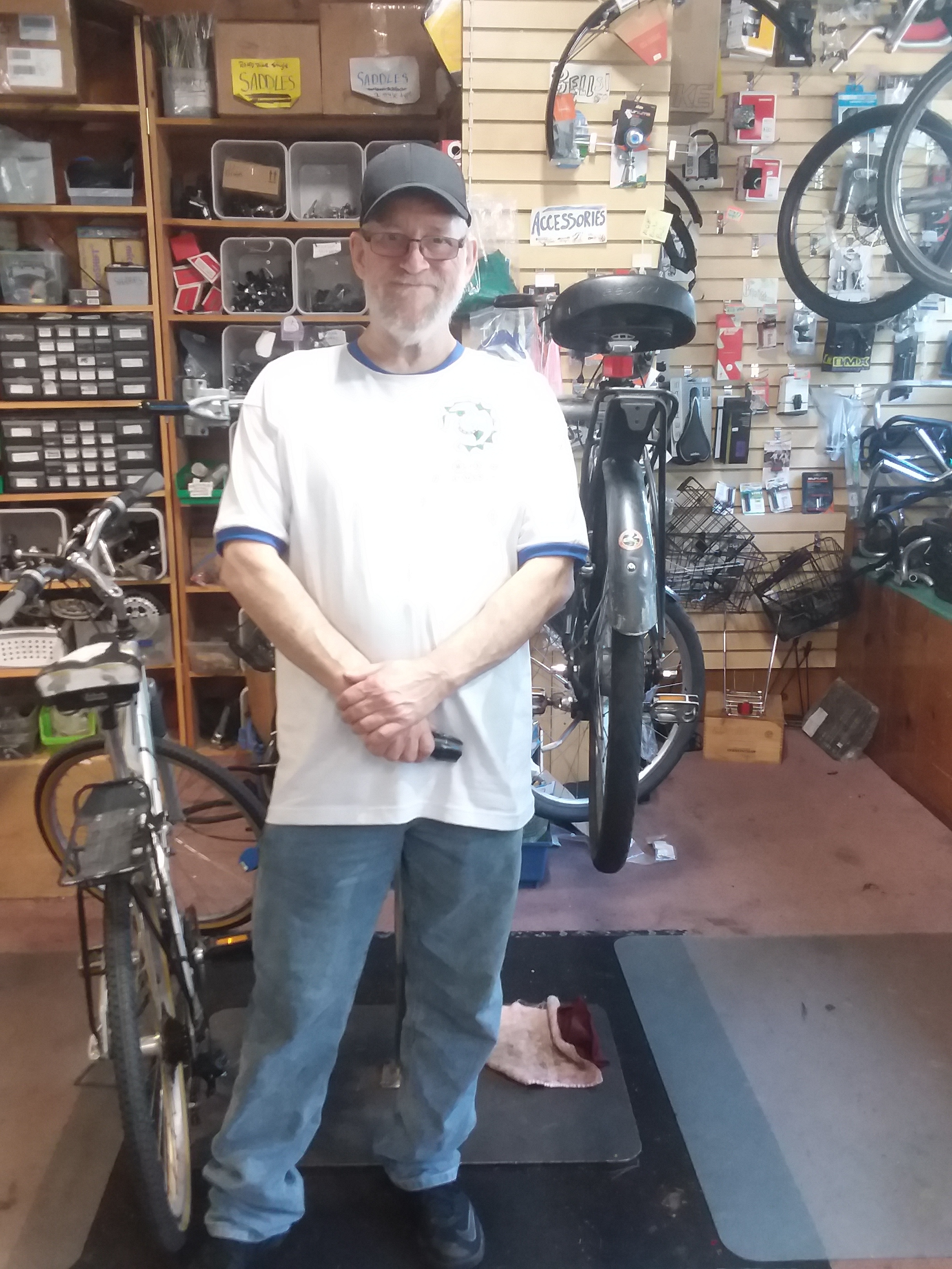 Image of Dave standing in the Bikes For Humanity PDX bike-shop in front of a bike stand. There is a bicycle to the right-hand side of the image and one suspended in the bike stand Dave is in front of.