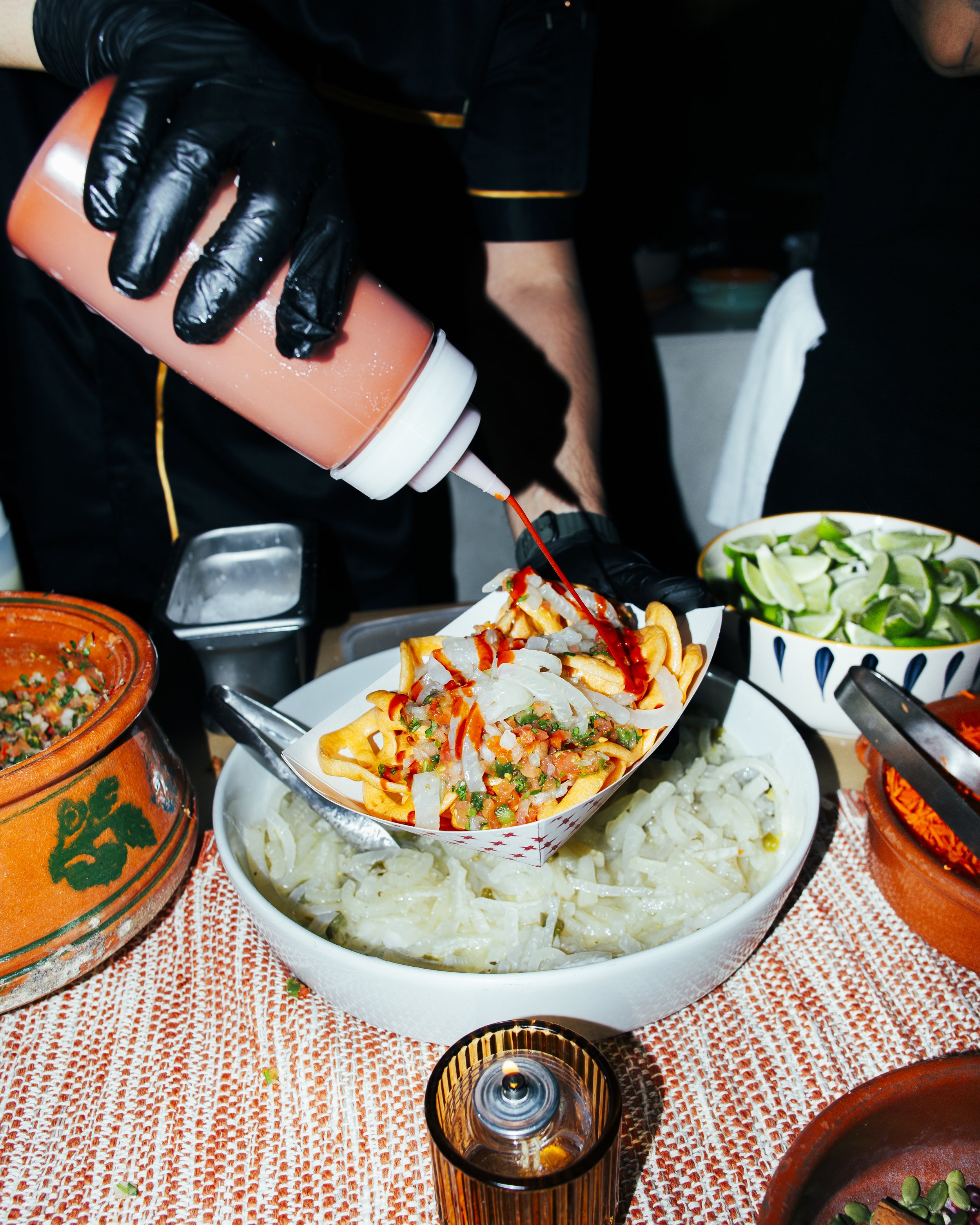 A person wearing black gloves is pouring a pink sauce from a squeeze bottle onto a plate of French fries topped with chili, onions, and peppers, placed over a bowl of cooked onions. Surrounding dishes include lime wedges, salsa, and various bowls of 