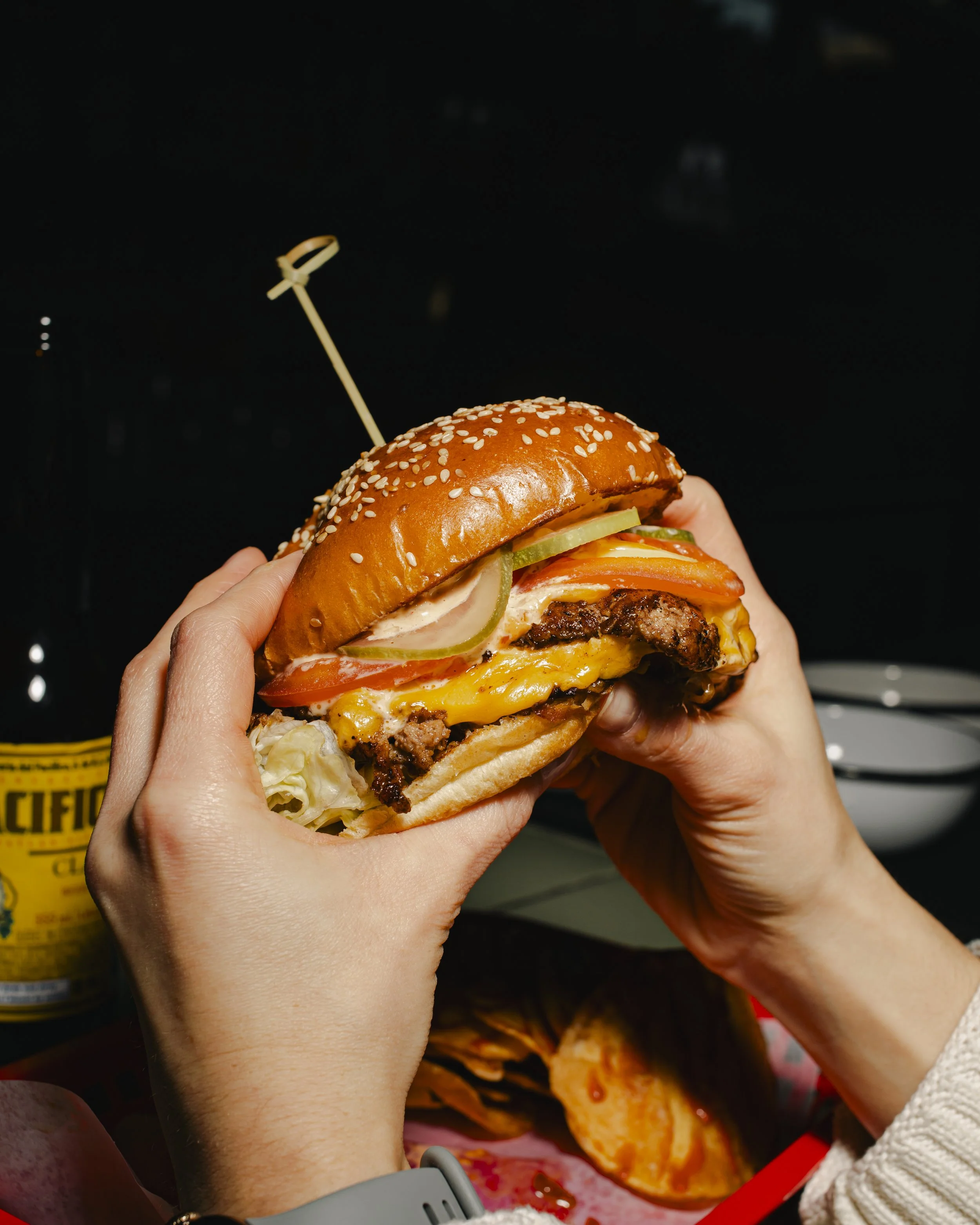 Person holding a large cheeseburger with lettuce, tomato, pickles, cheese, beef patty, and sesame seed bun