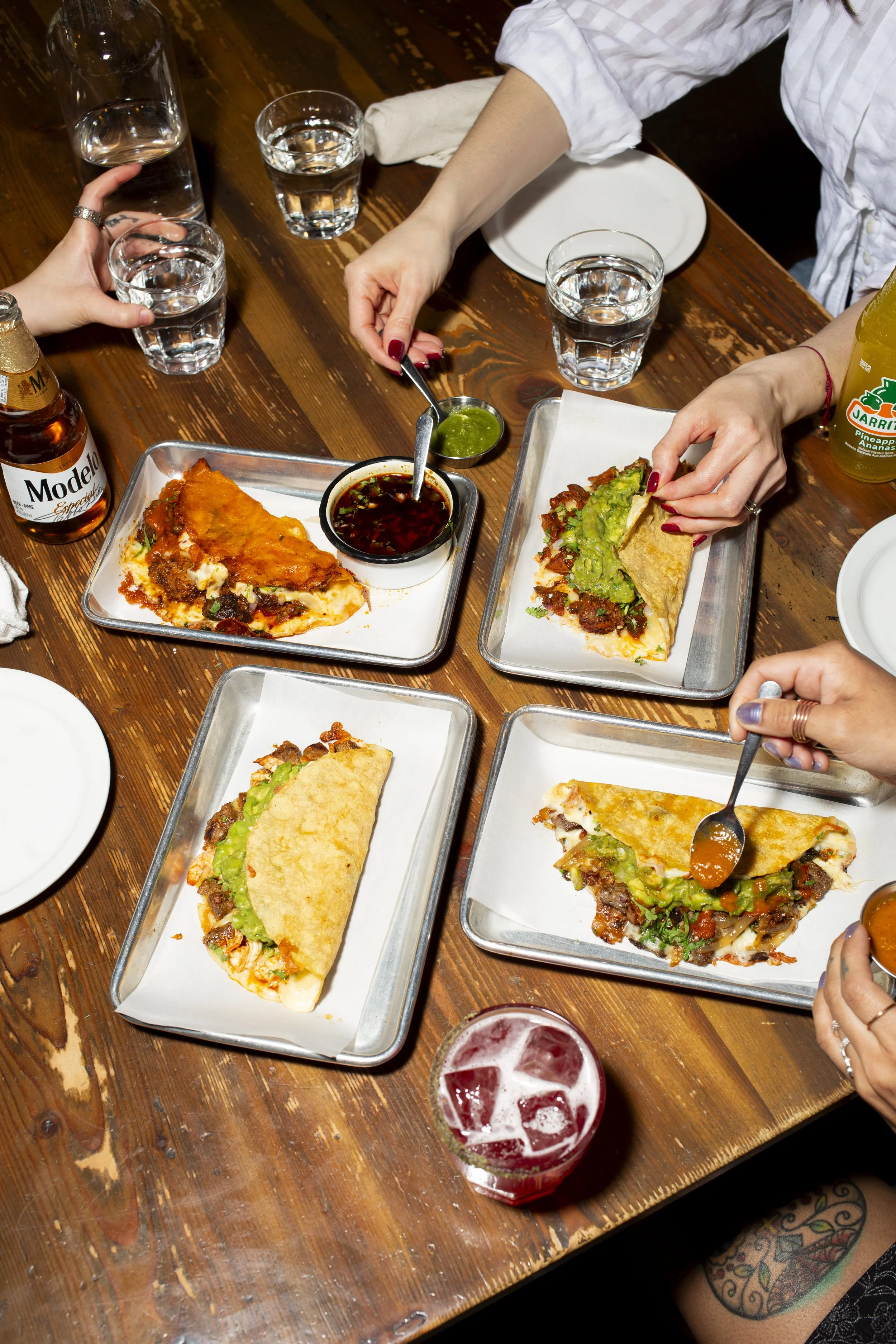 People enjoying Mexican food, including tacos and pizza, at a wooden table with drinks and dipping sauces.