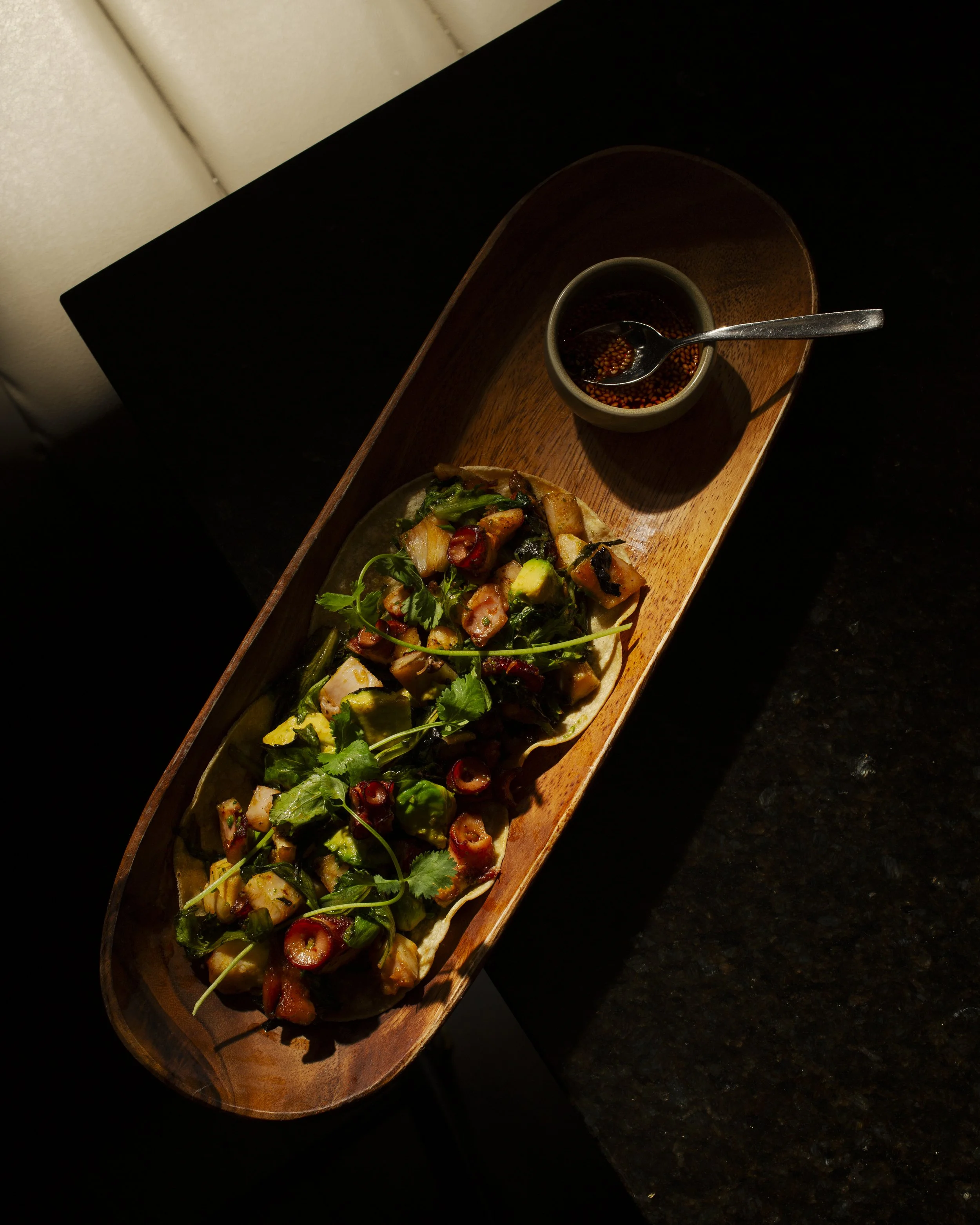 A long wooden serving board with a bowl of dipping sauce and a dish of chopped vegetables, herbs, and possibly seafood or meat, garnished with cilantro, on a dark countertop.
