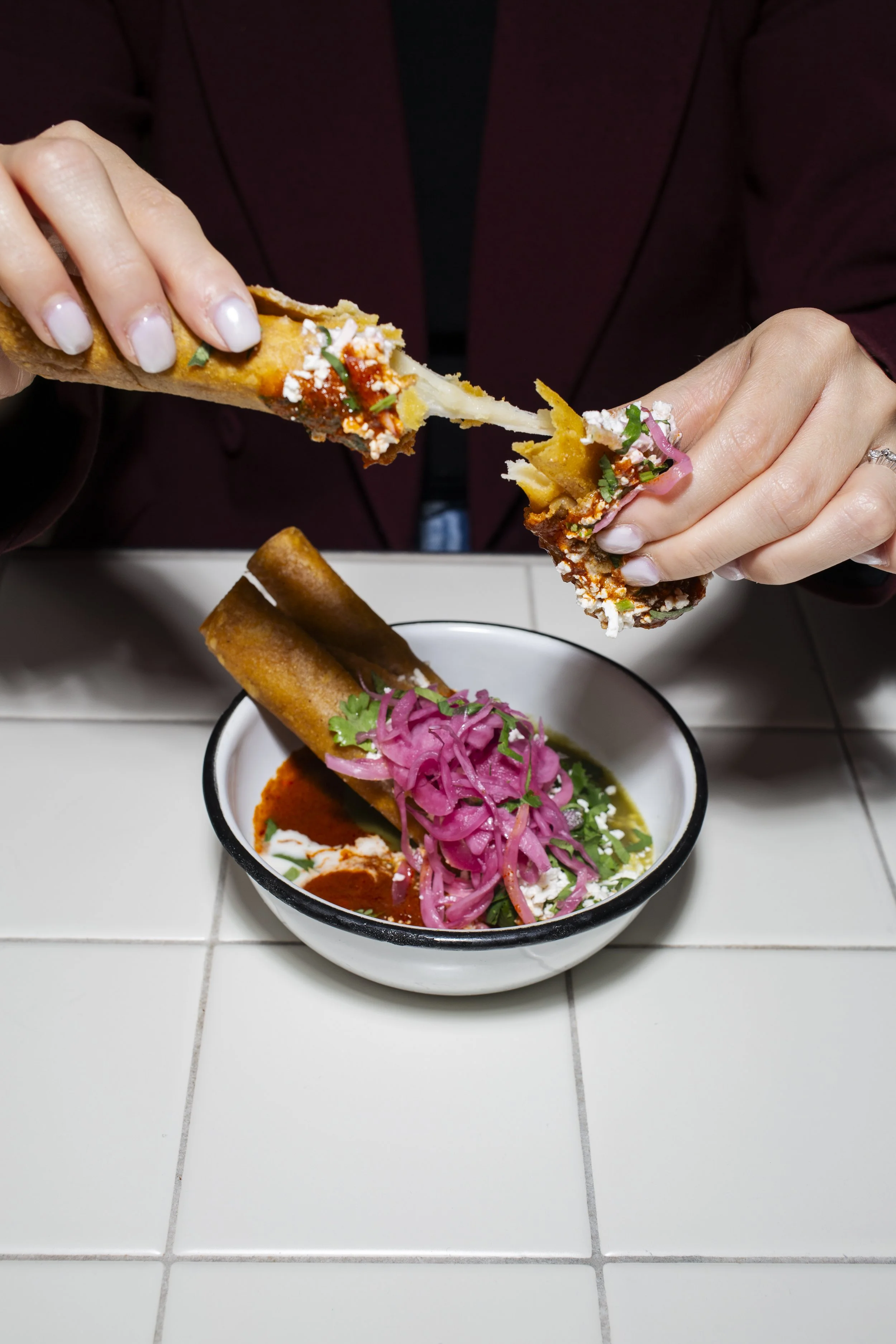 Person tearing apart a taquito with cheese and sauce over a bowl of Mexican soup garnished with pickled onions and cilantro on a tiled counter.